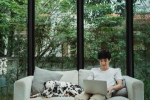 A man sits with a Dalmatian on a sofa, using a laptop, with a scenic outdoor view through large windows.